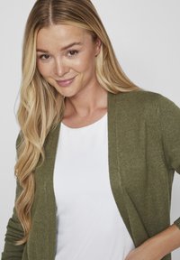 Green knitted cardigan with an open front, combined with a white top underneath. The texture appears soft and casual. Hair is long and wavy.