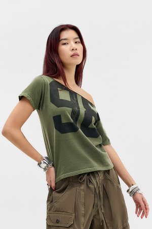 Giovane donna con lunghi capelli rossi che indossa una maglietta verde scollata con stampa nera e pantaloni cargo marroni larghi, posando su uno sfondo semplice.