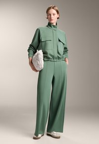 Completo sportivo verde a due pezzi con colletto alto, due tasche frontali e pantaloni a gamba larga, abbinato a una borsa grigia strutturata e scarpe da ginnastica.