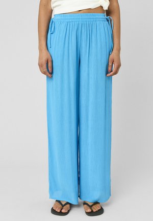 Femme portant un pantalon large ample bleu clair avec une taille élastique et des cordons, associé à des tongs noires sur un fond neutre.