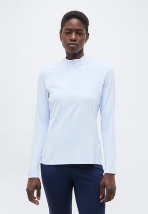 Naine, kes kannab helesinist pikkade varrukatega lukuga ülaosa ja tumelisi siniseid pükse, seisab ühevärvilise valge tausta ees.