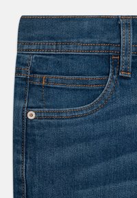 Σκούρες μπλε τζιν παντελόνες με μπροστινή τσέπη, εμφανή ραφή σε ανοιχτό πορτοκαλί και μεταλλικό πολιτιστικό διακοσμητικό στην άκρη της τσέπης.