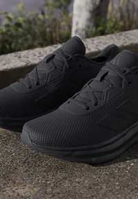 Paire de baskets noires de sport avec surface texturée et lacets, posées en extérieur sur du béton près d'un rebord en pierre et de plantes.