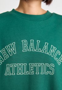 Felpa verde in cotone con scollatura a girocollo a costine. Presenta un testo stampato in bianco: “NEW BALANCE ATHLETICS” in grassetto, con un formato ad arco.