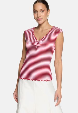 Femme portant un haut sans manches rayé rouge et blanc avec des bords festonnés et une jupe blanche, debout devant un fond uni.