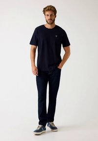 Granatowy bawełniany T-shirt z krótkim rękawem i małym logo na piersi, zestawiony z ciemnymi jeansami i niebieskimi trampkami z jasnymi akcentami.