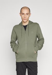 Olivinės žalios spalvos užtrauktukas su kišene, su raišteliu kapuce ir Lacoste logotipu. Lygus audinys, standartinė prigludimo forma ir ilgos rankovės.