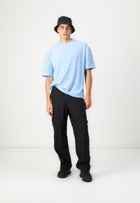 Lys blå teksturert t-skjorte med korte ermer, sammen med svarte cargo-bukser og svarte joggesko. Modellen har på seg en svart bøttehatt.