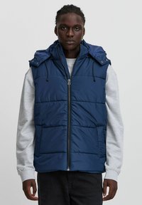 Tumši zila polsterēta veste ar priekšējo rāvējslēdzēju, regulējamu kapuci un divām priekšējām kabatām, izgatavota no viegla, gluda auduma.