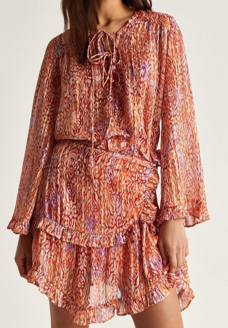 Femme portant une robe à manches longues, orange et rose à motifs, avec des détails à volants et une fermeture par lien au niveau de l'encolure.