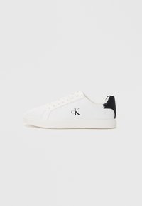 White low-top sneakers featuring a textured canvas upper, black heel accent, and a printed logo on the side. Flat rubber sole.
