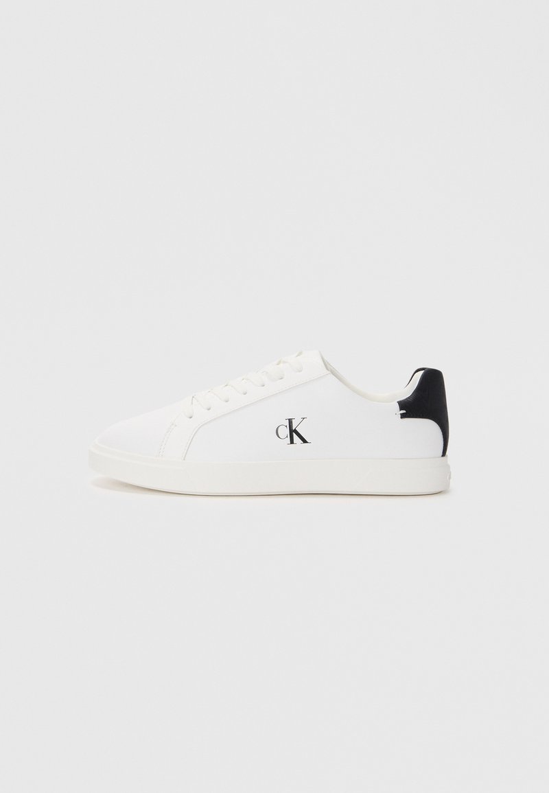 White low-top sneakers featuring a textured canvas upper, black heel accent, and a printed logo on the side. Flat rubber sole.