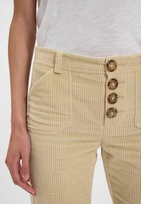 Pantalons en velours côtelé beige avec des côtes verticales, dotés d'une taille haute, de cinq boutons et de poches sur les côtés avant.
