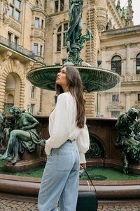 Jeune femme en chemisier blanc et jeans se tient près d'une fontaine ornée de statues en bronze vert dans une cour historique européenne.