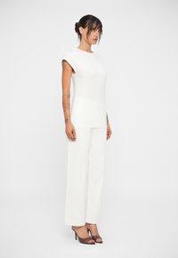 Haut blanc ajusté sans manches associé à un pantalon blanc ample. Texture lisse, design minimaliste, avec des chaussures à talons hauts ouvertes de couleur marron.