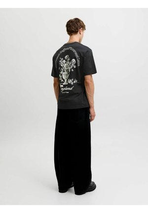 Ung mand med ryggen til iført en sort T-shirt med et hvidt blomsterkrukkeprint og tekst, kombineret med brede sorte bukser og sorte sko.