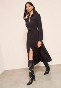 Must lõhikäistega must kleit, millel on krae, nööbid ees, vöökoht ja külgmine lõhik, koos mustade nahast sääriseidga.