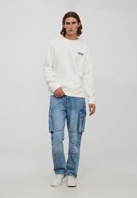 Biała bluza z czarnym napisem "VENICE", zestawiona z jasnoniebieskimi spodniami typu cargo z wieloma kieszeniami i luźnym krojem. Białe sneakersy dopełniają stylizację.