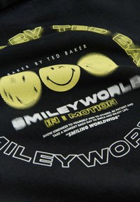 Tissu noir présentant des motifs de visages souriants jaunes et un texte en gras indiquant « SMILEYWORLD ». Comprend des motifs de points et des graphiques de chaque côté.