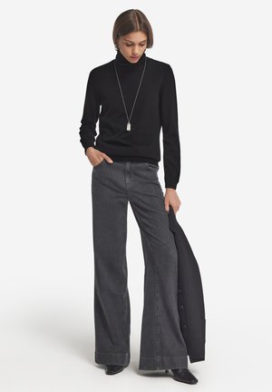 Schwarzer Rollkragenpullover, graue weite Jeans und schwarze spitze Stiefel. Auffällige silberne Anhängerkette und ein schwarzer Blazer über dem Arm.