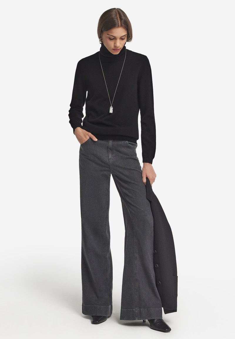 Schwarzer Rollkragenpullover, graue weite Jeans und schwarze spitze Stiefel. Auffällige silberne Anhängerkette und ein schwarzer Blazer über dem Arm.