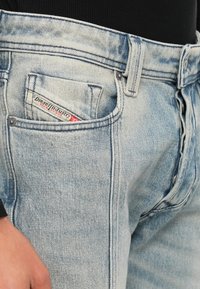 Φωτεινό μπλε τζιν με φθαρμένη υφή, που διαθέτει μια μικρή κόκκινη και λευκή ετικέτα στην μπροστινή τσέπη και μεταλλικά κουμπιά.