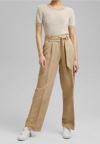 Beige korkeavyötäröiset housut, joissa on solmittava vyö ja laskostettu etuosa, yhdistettynä vaaleanbeigeen lyhythihaiseen neulepaitaan ja valkoisiin tennareihin.