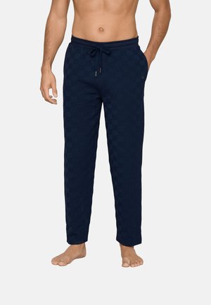 Man wearing navy blue checkered drawstring lounge pants standing barefoot with one hand in pocket and the other by his side.