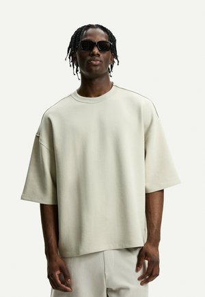 Hanorac oversized din bumbac bej deschis, cu mâneci scurte, gât rotund și detalii de cusătură subtile, acompaniat de pantaloni de culoare deschisă.