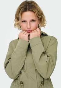 Veste à capuche vert olive avec taille à cordon de serrage, manches longues et détails de couture visibles. Texture lisse avec une coupe décontractée.
