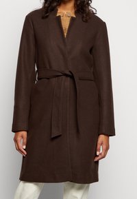 Donna che indossa un cappotto di lana marrone scuro con cintura su un maglione beige a bottoni e pantaloni bianchi, inquadrata dal collo alle ginocchia.
