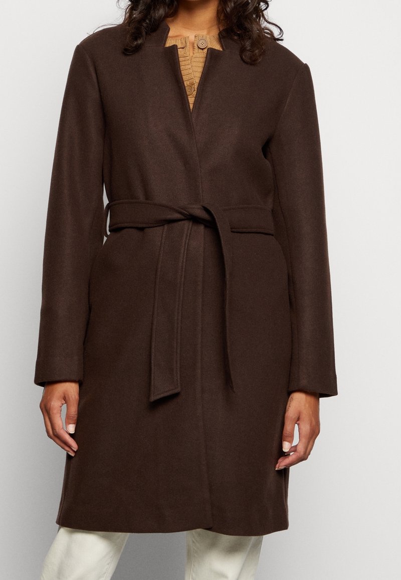 Donna che indossa un cappotto di lana marrone scuro con cintura su un maglione beige a bottoni e pantaloni bianchi, inquadrata dal collo alle ginocchia.