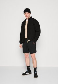 Black zip-up jacket, beige shirt, black shorts, black beanie. Black sneakers with white stripes and logo. Accessories include earrings.