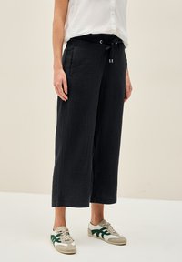 Una persona sta in piedi indossando una camicia chiara e pantaloni neri larghi e texturizzati. Indossa sneaker casual con accenti verdi.