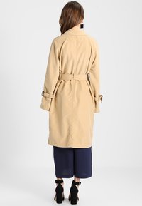 Trench-coat beige avec taille ceinturée, manches longues et poignets ajustables. Présente une texture lisse et des détails de coupe subtils.