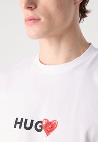 Balts kokvilnas T-krekls ar melnā krāsā uzrakstu "HUG" un sarkanu sirds dizainu. Apļveida dekoltē ar gludu tekstūru.