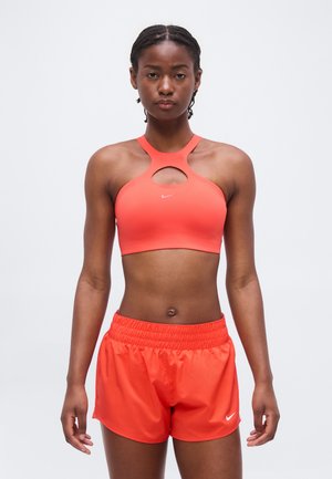 Atletyczna kobieta w jaskrawo pomarańczowym sportowym biustonoszu z wycięciem w kształcie klucza i pasujących szortach do biegania, stojąca na jednolitym tle.