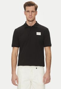 Maglietta polo nera con colletto, patta con tre bottoni e un piccolo etichetta bianca con il nome del marchio sul lato sinistro del petto.