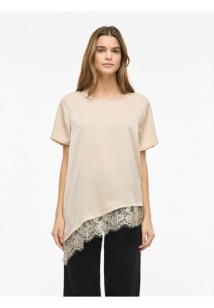 Kvinde iført beige kortærmet top med asymmetrisk kant og blonder, kombineret med sorte bukser, stående mod en hvid baggrund.