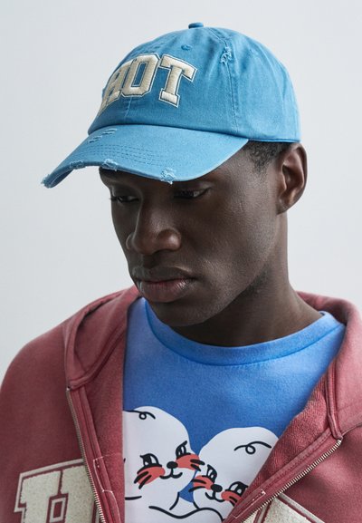 Gaiši zils džinsa cepure ar izšūtu vārdu "HOT" krēmkrāsā, ar nolietotām malām. Valkāta kopā ar sarkanu kapuci pār zilas grafiskās t-krekla attēlotām roņiem.