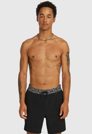 Muž bez trička s krátkymi kučeravými vlasmi a viacerými tetovaniami na rukách, oblečený v čiernych plavkových šortkách s vzorovaným pásom, stojaci na bielom pozadí.