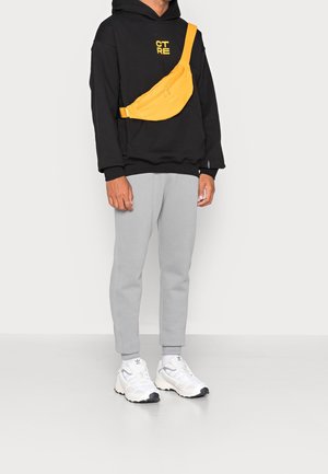 Bolso cruzado amarillo con textura suave, que cuenta con dos bolsillos con cremallera; llevado sobre un hoodie negro y pantalones de chándal grises, combinado con zapatillas blancas.