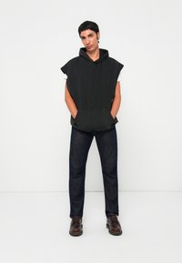 Schwarze Hoodie-Weste mit einer Fronttasche, oversize Form und kurzen Ärmeln, kombiniert mit dunklen Jeans und braunen Lederschuhen.