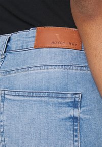 Ljusblå denimjeans med en jämn textur, med en brun läderetikett med präglad text "NOISY MAY". Inkluderar standard bakfickor.
