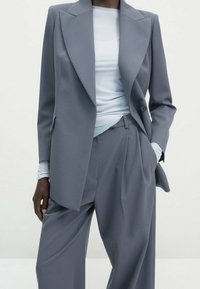 Lichtgrijze op maat gemaakte blazer met scherpe revers, dubbele knopen en relaxte fit broeken, laag over een aansluitend lichtblauw topje. Gladde textuur.