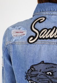 Ljusblå denimjacka med slitna lappar, med en stor svart broderad panterdesign och namnet "Saul" i krämfärgade bokstäver.