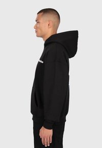 Man in een zwarte hoodie met een wit logo op de borst, staande in profiel tegen een effen lichte achtergrond.