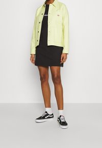 Ljusgul, knappad jacka med bröstficka över en svart T-shirtklänning. Matchas med svarta sneakers och vita ankelstrumpor.