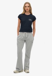 Vrouw die recht vooruit staat, gekleed in een zwart Superdry t-shirt, grijze sweatpants met trekkoord en beige sneakers op een witte achtergrond.