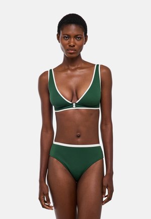 Femme portant un bikini vert foncé avec bordure blanche, debout face à un fond uni et clair.